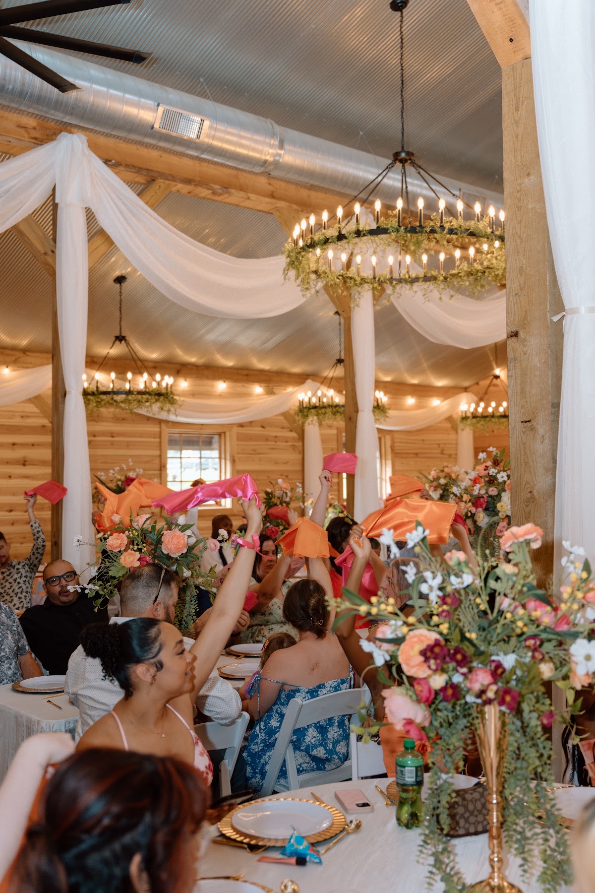 Guests wave bright pink and orange napkins beneath greenery chandeliers for Stephanie & Josh at Covey Creek
