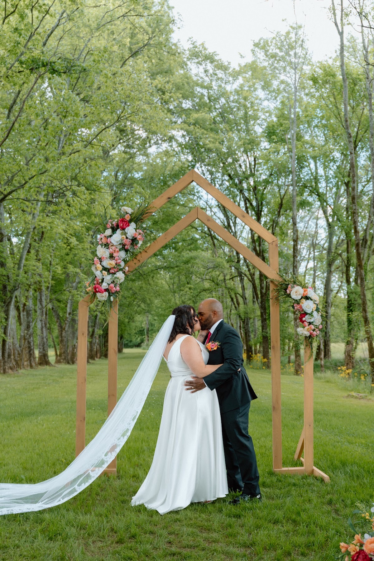 Stephanie & Josh kiss under a floral wooden arch, her long white veil trailing on the grass at Covey Creek