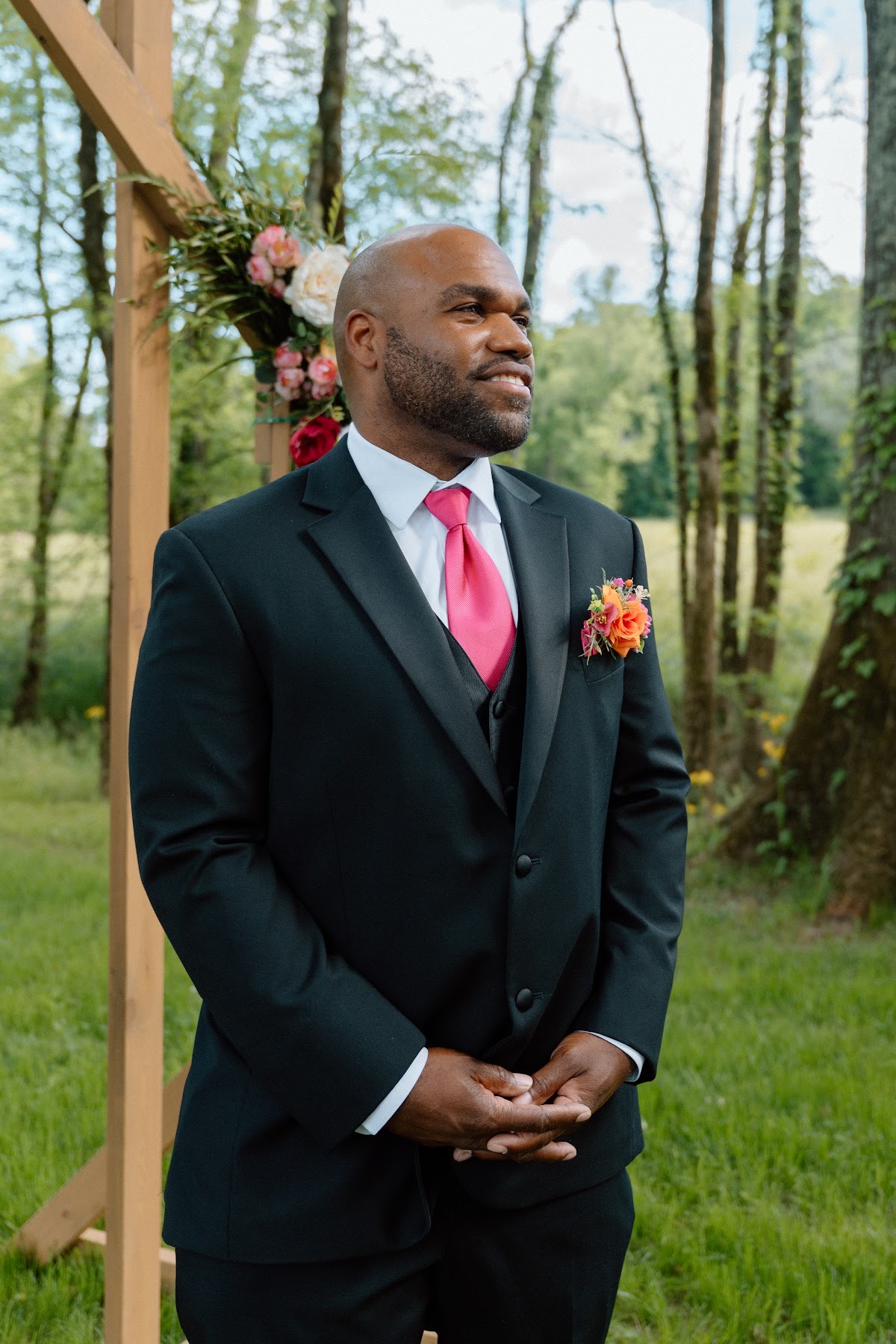 Josh smiles in a black suit and bright pink tie beside the floral wooden arch for Stephanie & Josh at Covey Creek