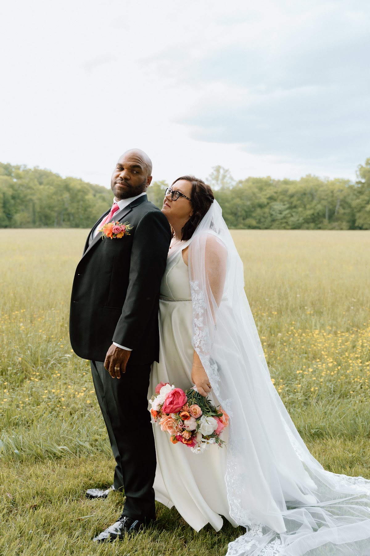 Stephanie & Josh stand back-to-back in a green field, her pink bouquet and his pink tie at Covey Creek