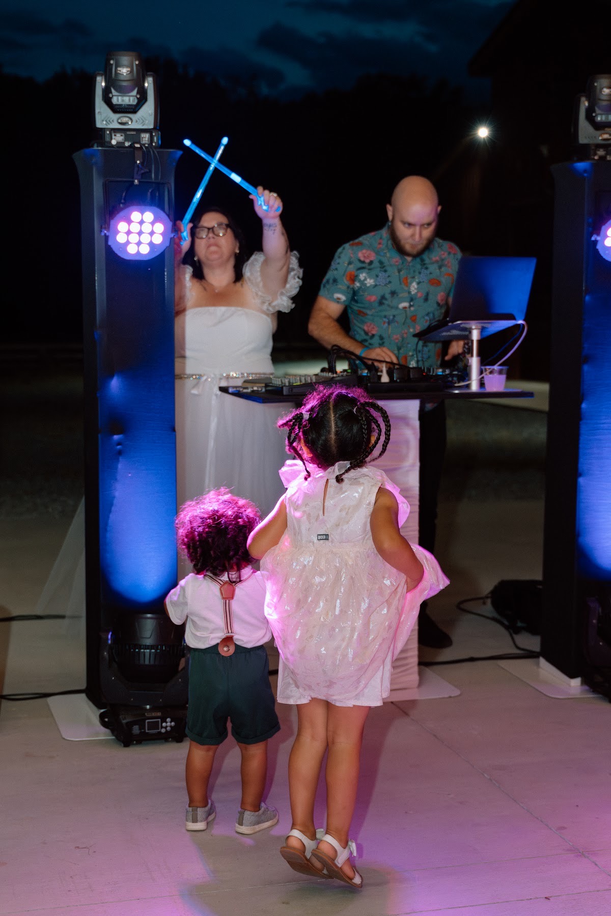 Stephanie raises glowing blue sticks at the DJ booth as children watch for Stephanie & Josh at Covey Creek