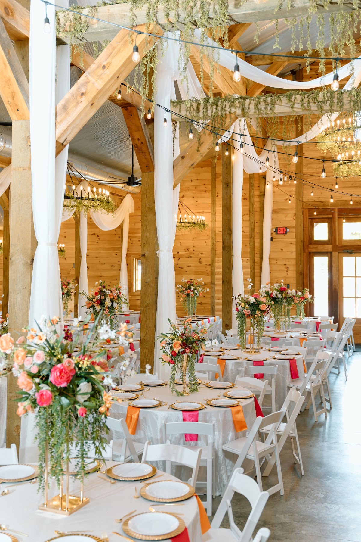 Round tables with tall gold florals and pink-orange napkins fill the barn for Stephanie & Josh at Covey Creek