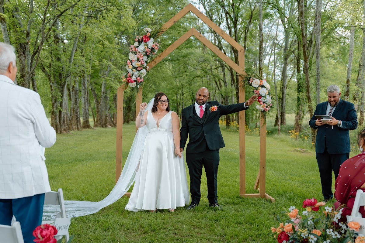 Stephanie raises a fist as she and Josh celebrate under the floral wooden arch at Covey Creek