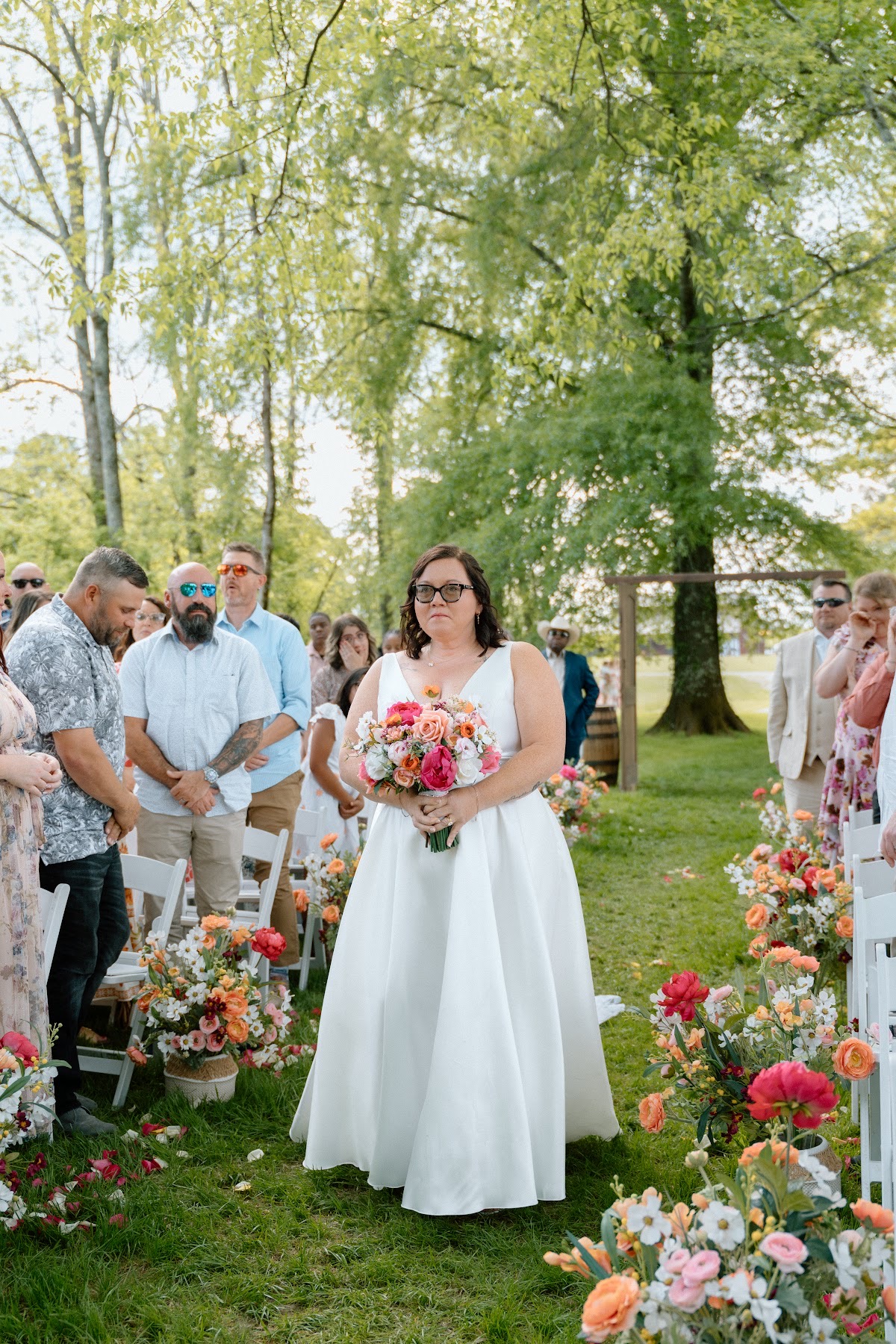 Stephanie walks the grassy aisle with a pink and orange bouquet toward Josh at Covey Creek