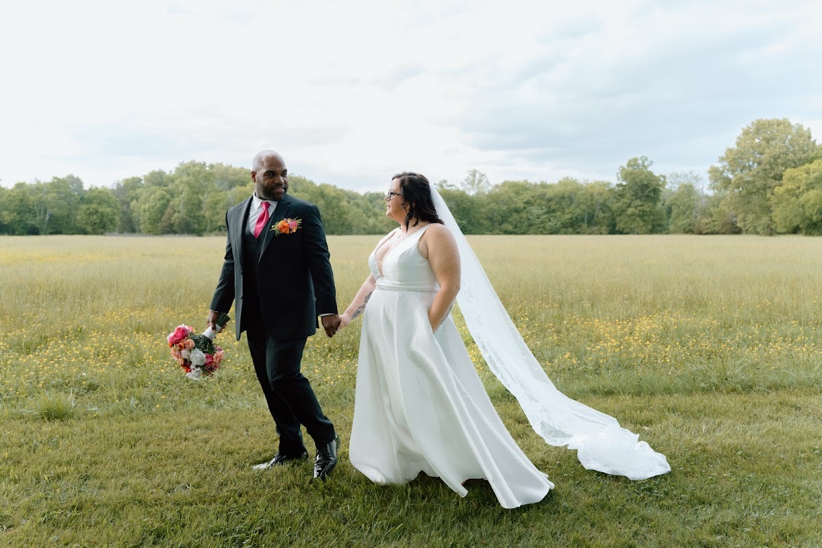Stephanie & Josh walk hand in hand across a wildflower meadow, her veil trailing at Covey Creek