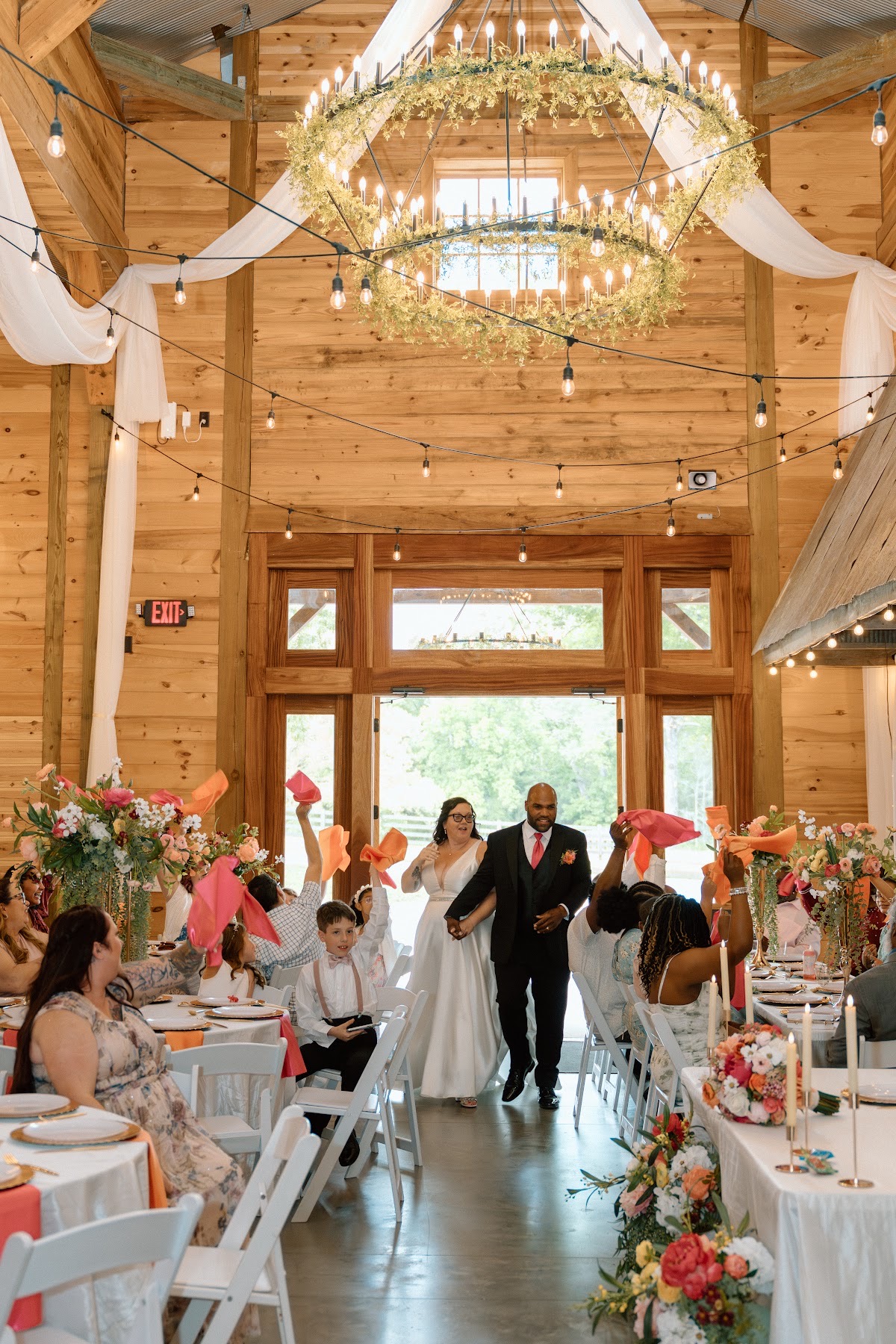 Stephanie & Josh enter the barn hand in hand as guests wave pink and orange napkins at Covey Creek